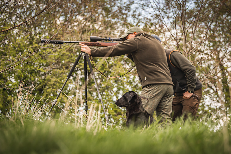 two men mounting rifle in woods to stalk deer, with a black Labrador by their side
